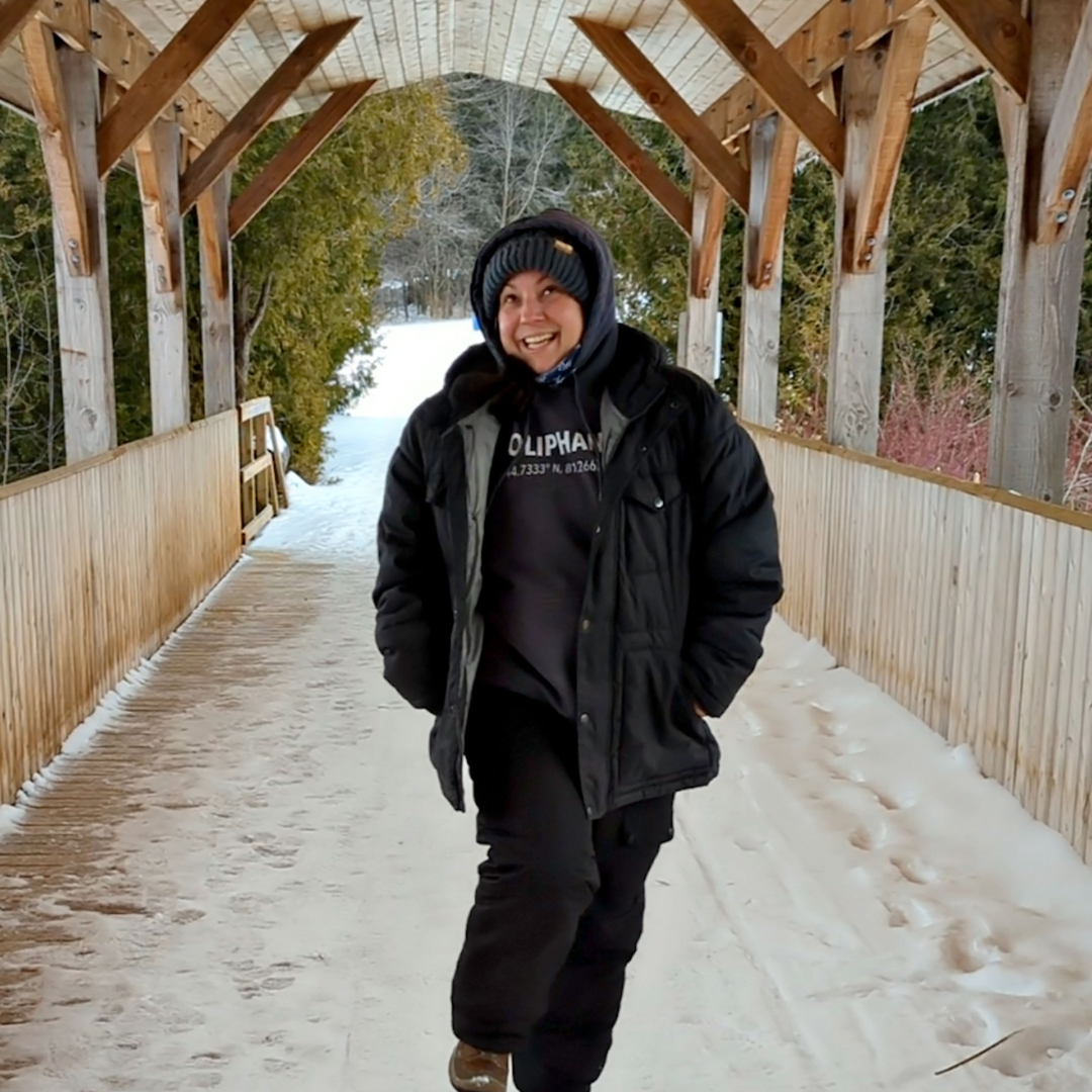 A smiling woman in winter apparel, stands on a covered bridge with snow on the ground.